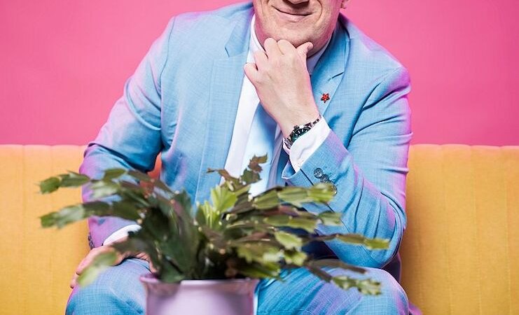 Ein Mann in einem blauen Anzug sitzt l&auml;chelnd auf einem gelben Sofa vor einem pinken Hintergrund. Eine Pflanze in einem lila Topf steht auf einem Tisch vor ihm., &copy; Stadthalle Leonberg