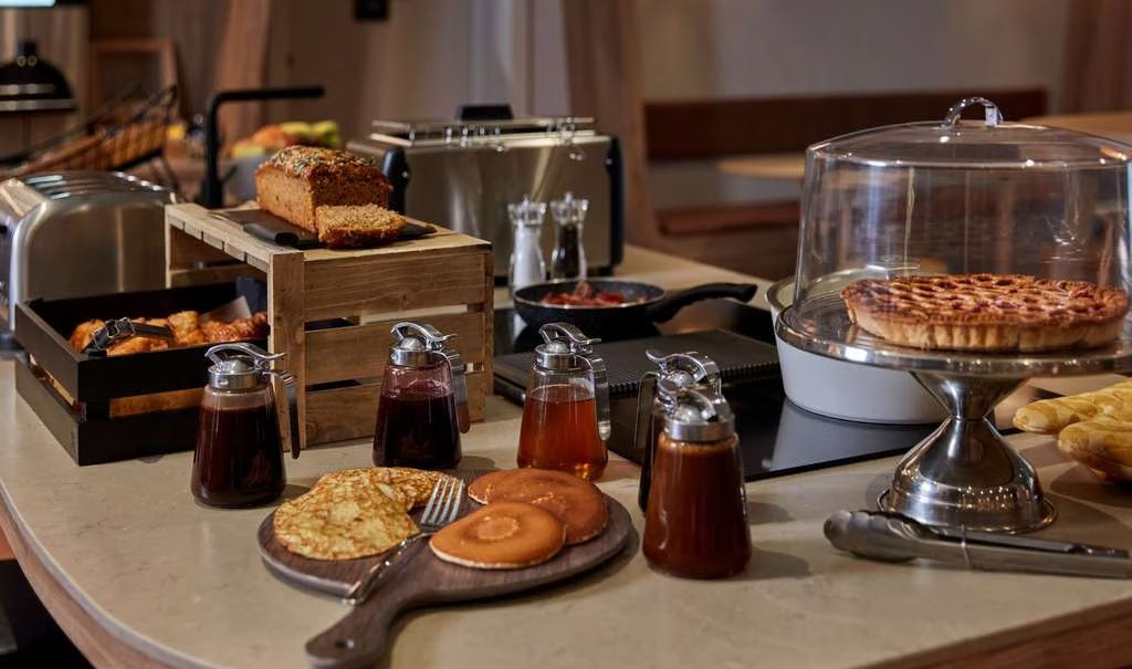 Ein Frühstücksbuffet mit verschiedenen Gebäcksorten, Sirupflaschen, Brot und einem Kuchen unter einer Glasglocke auf einem Tisch., © Adagio Ein Frühstücksbuffet mit verschiedenen Gebäcksorten, Sirupflaschen, Brot und einem Kuchen unter einer Glasglocke auf einem Tisch., © Adagio