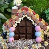 Ein mit Schokolade und bunten Süßigkeiten dekoriertes Lebkuchenhaus vor einem Hintergrund aus grünen Blättern., © Alfred Ritter GmbH &amp; Co. KG | Waldenbuch