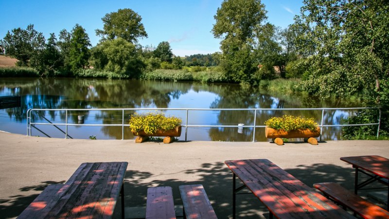 Ein idyllischer See mit B&auml;umen im Hintergrund, umgeben von Natur. Im Vordergrund stehen B&auml;nke und Blumenk&auml;sten mit gelben Blumen., &copy; agentur arcos/Niki Eilers