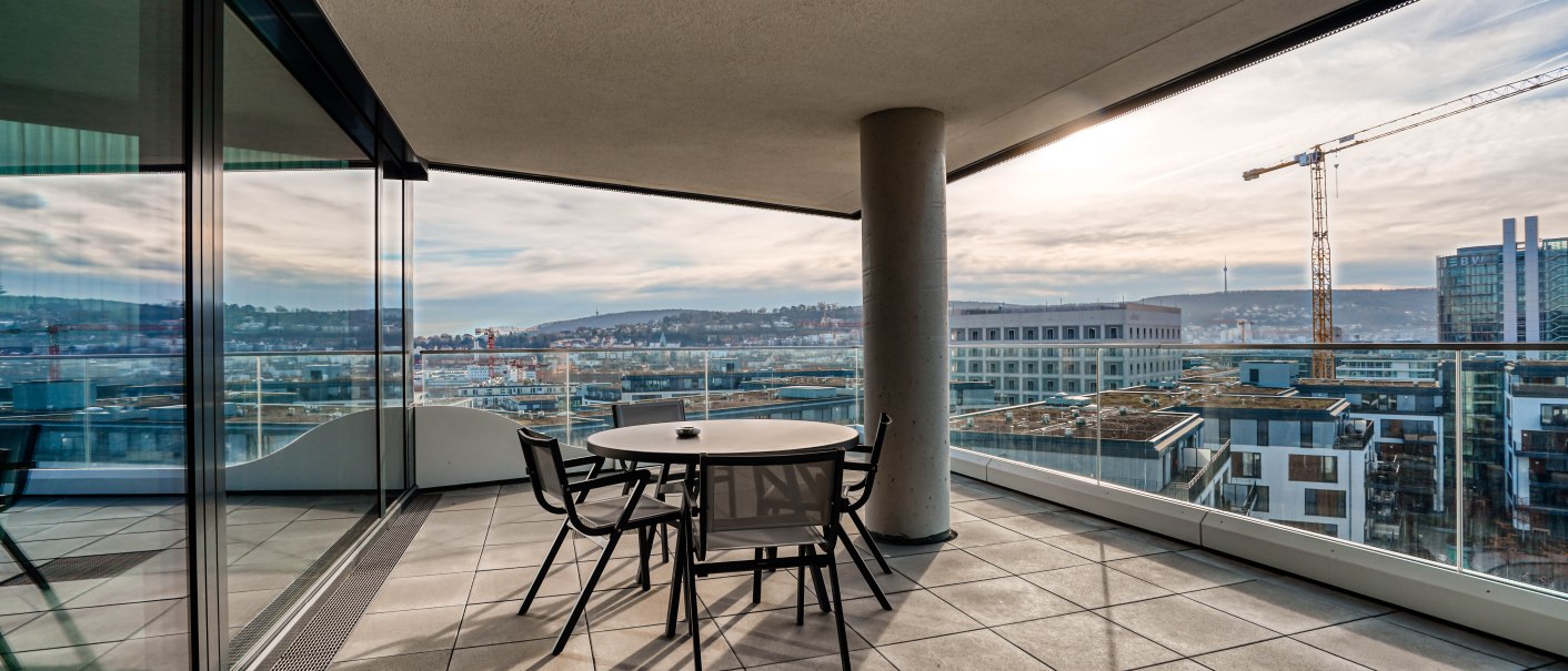 Moderner Balkon mit Tisch und Stühlen, bietet einen weiten Blick über die Stadt und einen Baukran in der Ferne., © CLOUD N°7 APARTMENTS GmbH