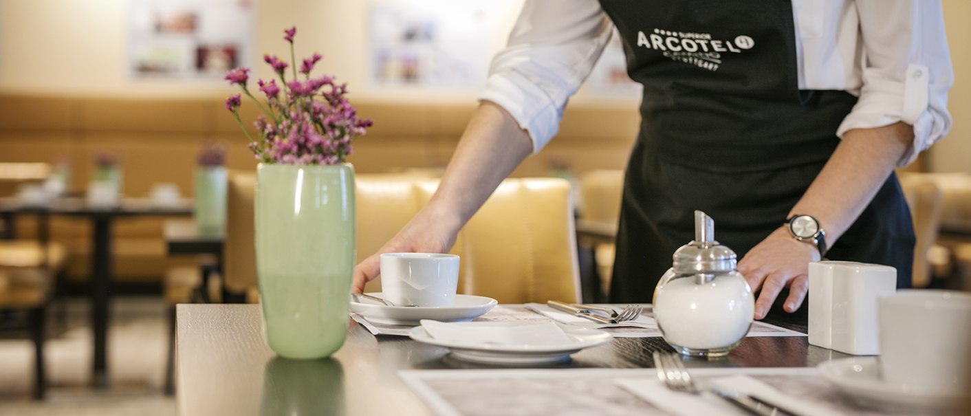 Eine Person deckt einen Tisch in einem Restaurant. Auf dem Tisch stehen eine Tasse, eine Zuckerdose und eine Vase mit Blumen., &copy; Arcotel Hotels