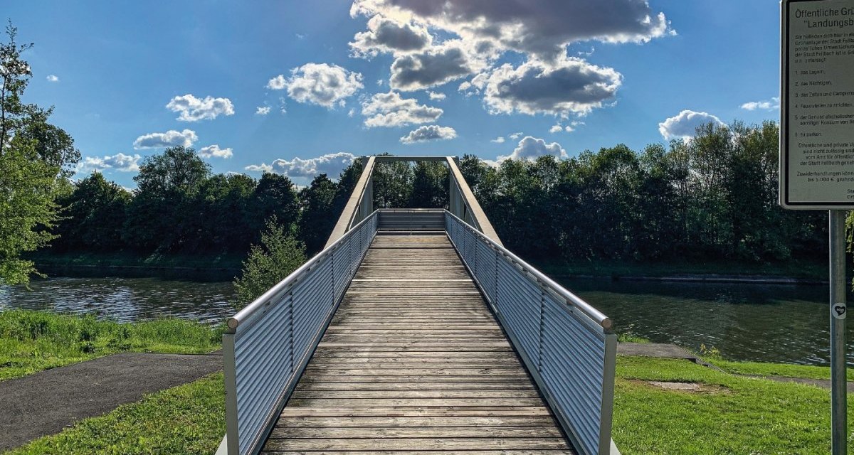 Eine Holzbrücke führt über einen Fluss, umgeben von grüner Landschaft. Der Himmel ist blau mit einigen Wolken, die Sonne scheint hell., © Remstal Tourismus e.V. Eine Holzbrücke führt über einen Fluss, umgeben von grüner Landschaft. Der Himmel ist blau mit einigen Wolken, die Sonne scheint hell., © Remstal Tourismus e.V.