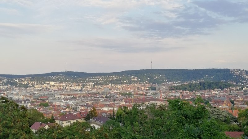 Blick über eine Stadt mit einem Fernsehturm in der Ferne, umgeben von Hügeln und bewaldeten Flächen im Vordergrund., © SMG
