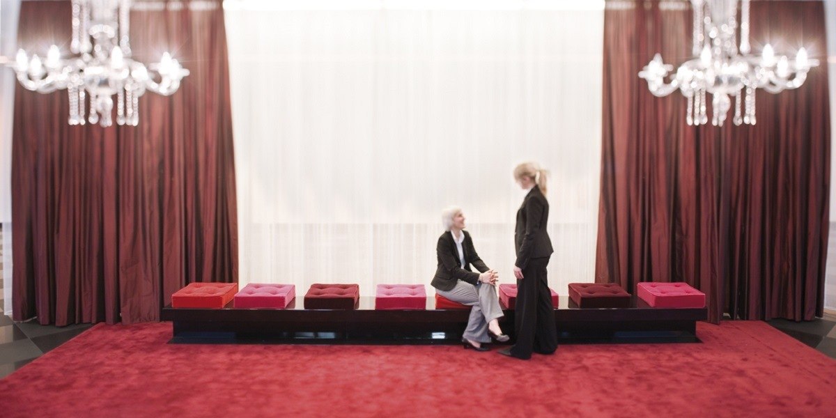 Zwei Personen in einer eleganten Lobby mit roten Vorhängen, Kronleuchtern und einer Sitzbank mit bunten Kissen., © Mövenpick Hotel Stuttgart Airport