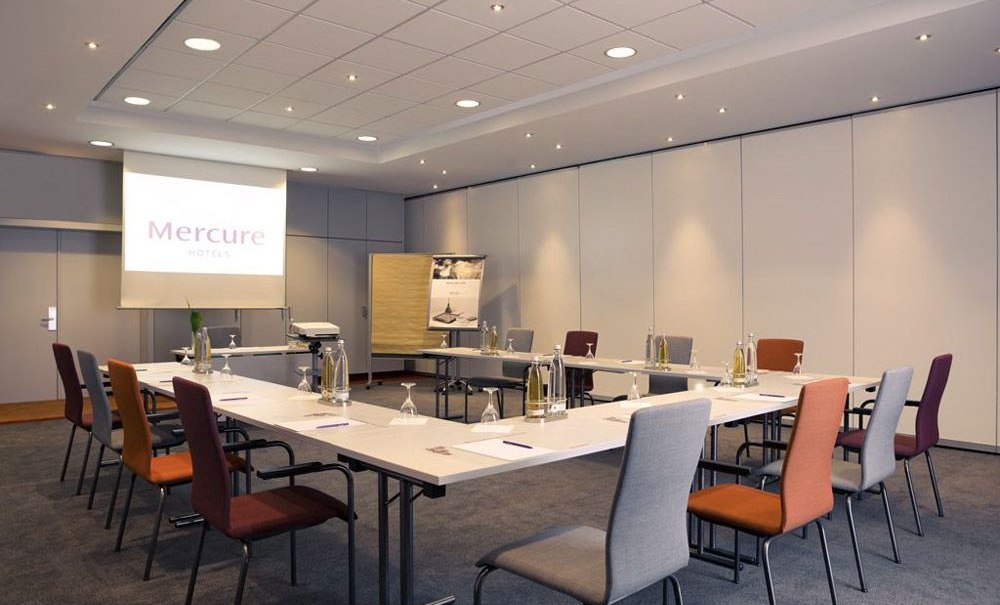 Moderner Tagungsraum mit U-förmiger Tischanordnung, Stühlen in verschiedenen Farben und Präsentationstechnik. An der Wand ein Projektorbild mit 'Mercure Hotels'., © Mercure Stuttgart Airport Messe Moderner Tagungsraum mit U-förmiger Tischanordnung, Stühlen in verschiedenen Farben und Präsentationstechnik. An der Wand ein Projektorbild mit 'Mercure Hotels'., © Mercure Stuttgart Airport Messe