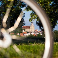 Ein Fahrrad im Vordergrund, durch dessen Speichen eine Kirche in einer Stadt zu sehen ist, umgeben von Bäumen und grüner Landschaft., © Natur.Nah. Schönbuch & Heckengäu