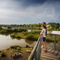 Zwei Personen auf einer Plattform blicken auf den Neckar und die grüne Landschaft. Eine Person liegt auf einer Bank am Flussufer. Himmel ist klar., © Stoppel