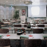 Ein leerer Konferenzraum mit Tischen, Stühlen, einem Projektor und einem Flipchart. Auf den Tischen liegen Ordner und Wasserflaschen., © © H-Hotels.com