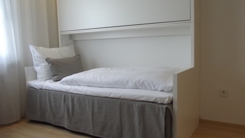 Ein Einzelbett mit weißer Bettwäsche und grauem Rock steht in einem Zimmer. Ein Etagenbett ist in die Wand eingeklappt., © Hotel am Engelberg