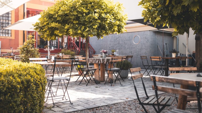Ein leerer Café-Außenbereich mit Holztischen und -stühlen, umgeben von grünen Bäumen und Pflanzen, auf einem gepflasterten Boden., © © N. Trautmann