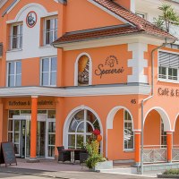 Orangefarbenes Gebäude mit Aufschrift 'Kaffeehaus-Spezerei Sanct Bernhard'. Menschen sitzen draußen an Tischen. Fahrräder stehen vor dem Gebäude., © Kräuterhaus Sanct Bernhard KG