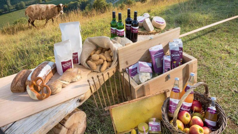Regionale Produkte wie Brot, Wein, Äpfel und Honig auf einem Tisch im Freien. Ein Schaf steht im Hintergrund auf einer Wiese. Regionale Produkte wie Brot, Wein, Äpfel und Honig auf einem Tisch im Freien. Ein Schaf steht im Hintergrund auf einer Wiese.