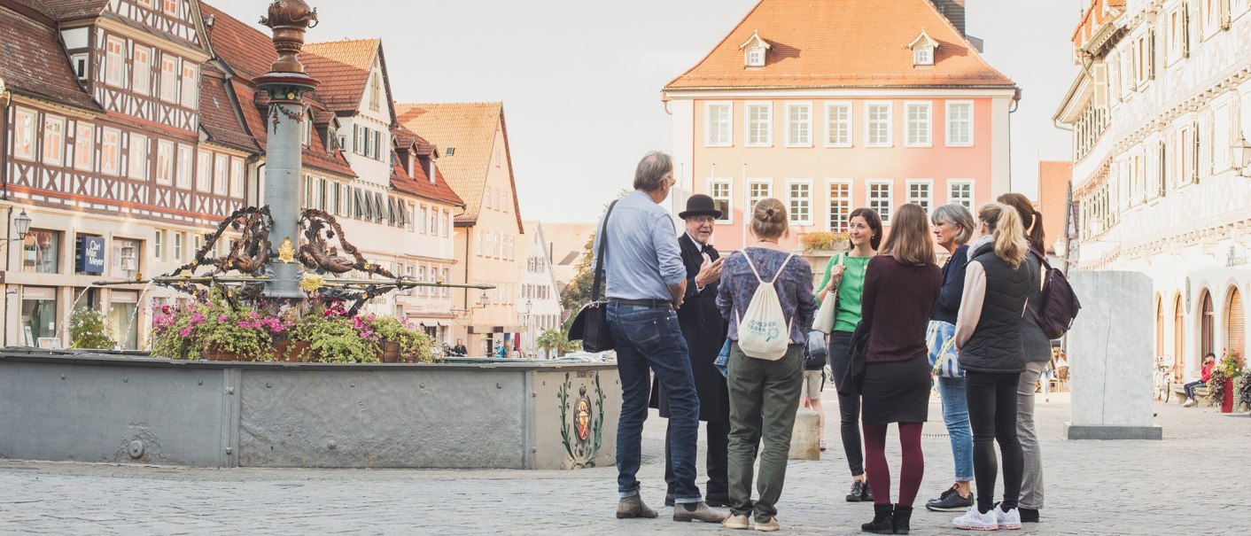 Eine Gruppe von Menschen steht um einen Marktbrunnen in einer Altstadt mit Fachwerkhäusern. Ein Mann in Hut und Mantel scheint eine Führung zu geben., © Bebop Media, Danijel Grbic Eine Gruppe von Menschen steht um einen Marktbrunnen in einer Altstadt mit Fachwerkhäusern. Ein Mann in Hut und Mantel scheint eine Führung zu geben., © Bebop Media, Danijel Grbic
