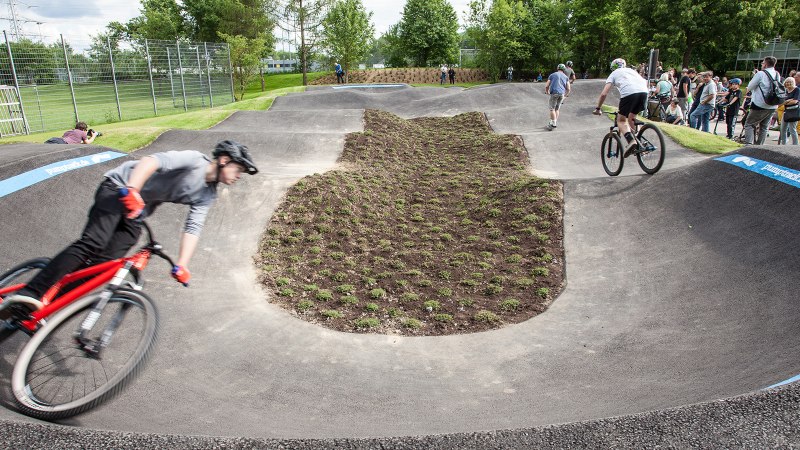 Fahrradfahrer auf einem Pumptrack in Sindelfingen, umgeben von Zuschauern und Bäumen. Die Strecke ist asphaltiert und wellig., © Fotoknobi Fahrradfahrer auf einem Pumptrack in Sindelfingen, umgeben von Zuschauern und Bäumen. Die Strecke ist asphaltiert und wellig., © Fotoknobi