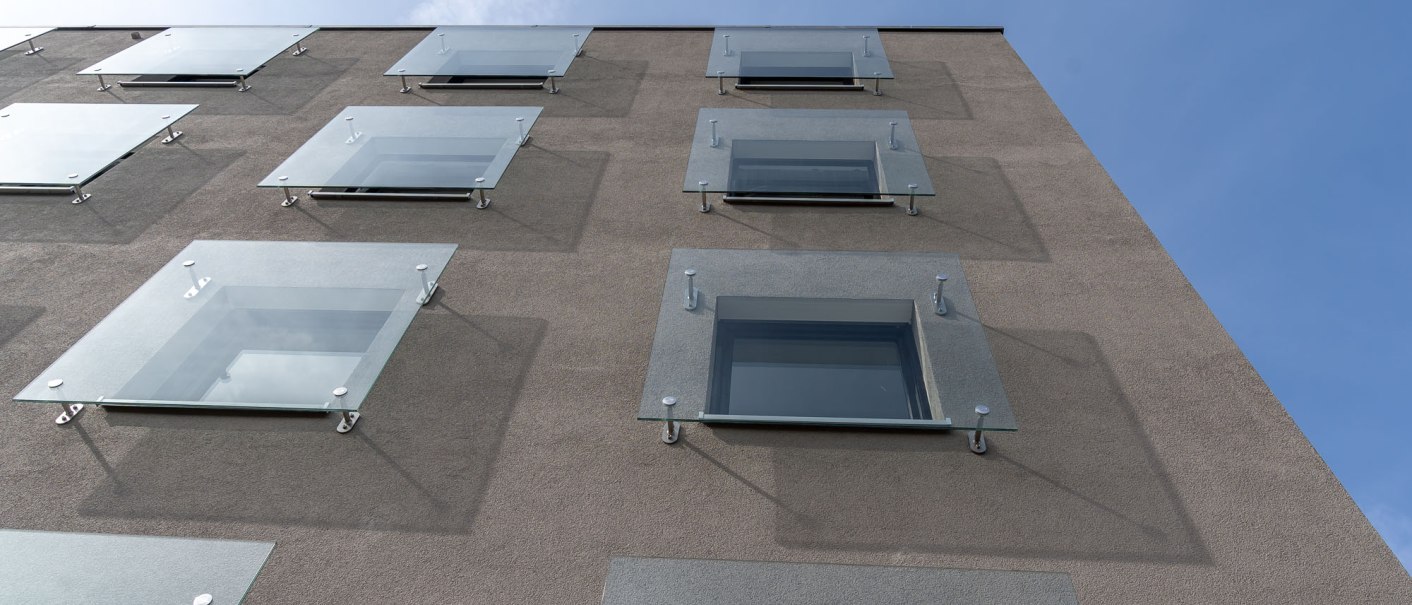 Moderne Gebäudefassade mit Glasvordächern über den Fenstern, die in einem geometrischen Muster angeordnet sind. Blauer Himmel im Hintergrund., © a2 HOTELS