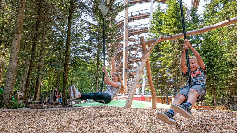Zwei Kinder schaukeln fröhlich im Abenteuerwald Sommerberg. Im Hintergrund ist ein großer Holzpfad zu sehen, umgeben von hohen Bäumen., © Erlebnis Akademie AG Zwei Kinder schaukeln fröhlich im Abenteuerwald Sommerberg. Im Hintergrund ist ein großer Holzpfad zu sehen, umgeben von hohen Bäumen., © Erlebnis Akademie AG