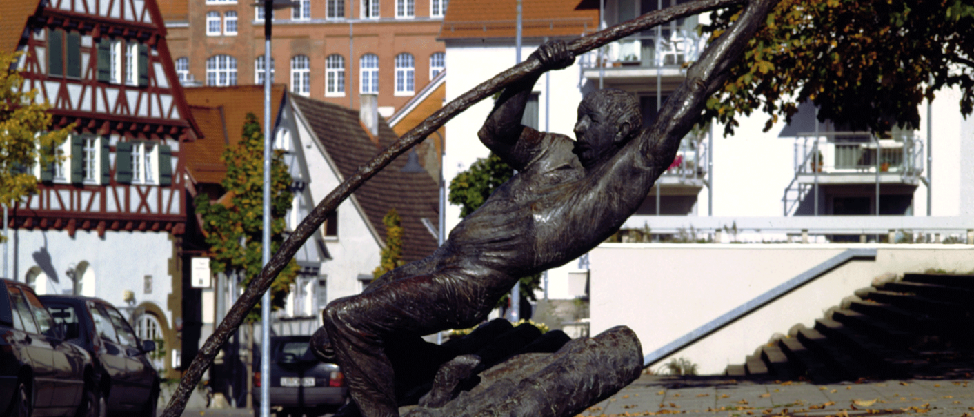 Bronzestatue eines Flößers, der mit einem langen Stab auf einem Baumstamm balanciert. Im Hintergrund Fachwerkhäuser und Herbstlaub auf dem Boden., © Stuttgart-Marketing GmbH