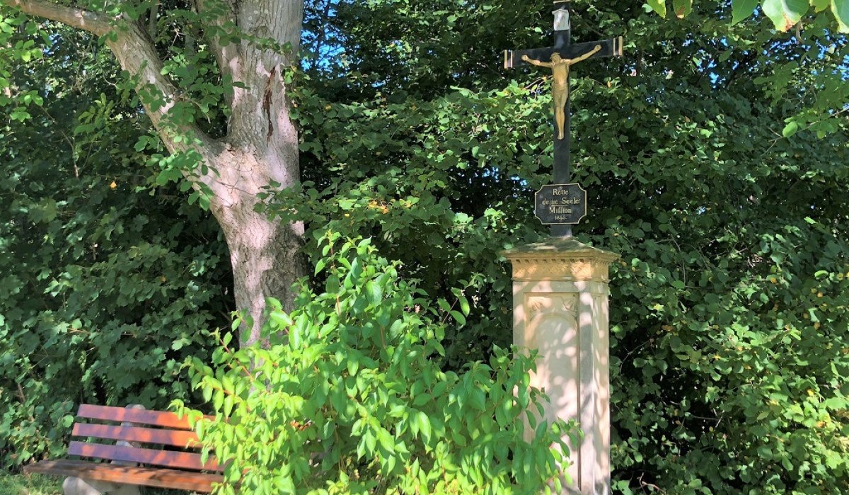 Ein steinernes Kreuz mit Jesusfigur steht neben einer Holzbank unter einem Baum, umgeben von dichtem Grün., © Touristik und Marketing GmbH Ein steinernes Kreuz mit Jesusfigur steht neben einer Holzbank unter einem Baum, umgeben von dichtem Grün., © Touristik und Marketing GmbH
