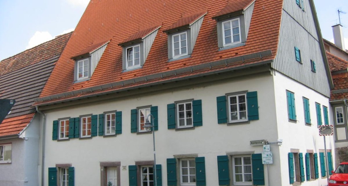 Ein zweistöckiges Bauernhaus mit roten Dachziegeln und grünen Fensterläden. Es steht an einer Straßenecke mit einem Straßenschild und geparkten Autos., © Land der 1000 Hügel - Kraichgau-Stromberg Ein zweistöckiges Bauernhaus mit roten Dachziegeln und grünen Fensterläden. Es steht an einer Straßenecke mit einem Straßenschild und geparkten Autos., © Land der 1000 Hügel - Kraichgau-Stromberg
