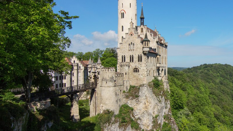 Schloss Lichtenstein thront auf einem Felsen, verbunden durch eine Brücke, umgeben von üppigem Wald und blauem Himmel., © Armin Dieter