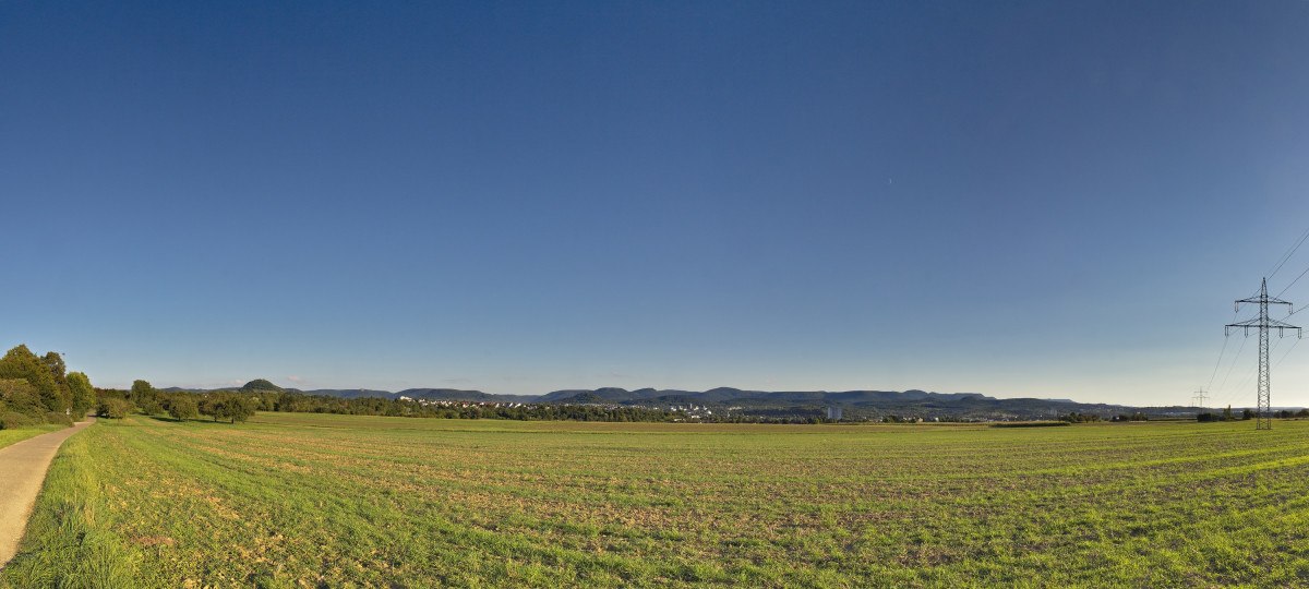 Weite Landschaft mit Feldern, einem Weg links, Strommasten rechts und einem klaren blauen Himmel. Im Hintergrund sind Hügel zu sehen., © Mythos Schwäbische Alb Weite Landschaft mit Feldern, einem Weg links, Strommasten rechts und einem klaren blauen Himmel. Im Hintergrund sind Hügel zu sehen., © Mythos Schwäbische Alb