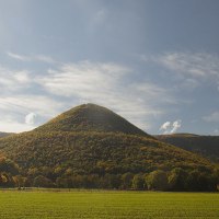 Ein runder, bewaldeter Berg erhebt sich unter einem blauen Himmel mit Wolken. Im Vordergrund sind grüne Felder und Bäume zu sehen., © Bad Urach Tourismus Ein runder, bewaldeter Berg erhebt sich unter einem blauen Himmel mit Wolken. Im Vordergrund sind grüne Felder und Bäume zu sehen., © Bad Urach Tourismus