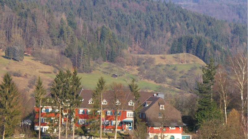Ein rotes Gebäude mit weißen Fenstern steht vor einem bewaldeten Hügel. Im Vordergrund sind Bäume und Sträucher zu sehen., © DJH Baden-Württemberg e.V.