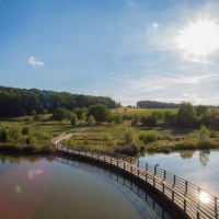 Holzsteg &uuml;ber einen Teich in einer gr&uuml;nen Landschaft mit H&uuml;geln und B&auml;umen, unter strahlendem Sonnenschein., &copy; &copy; T&E