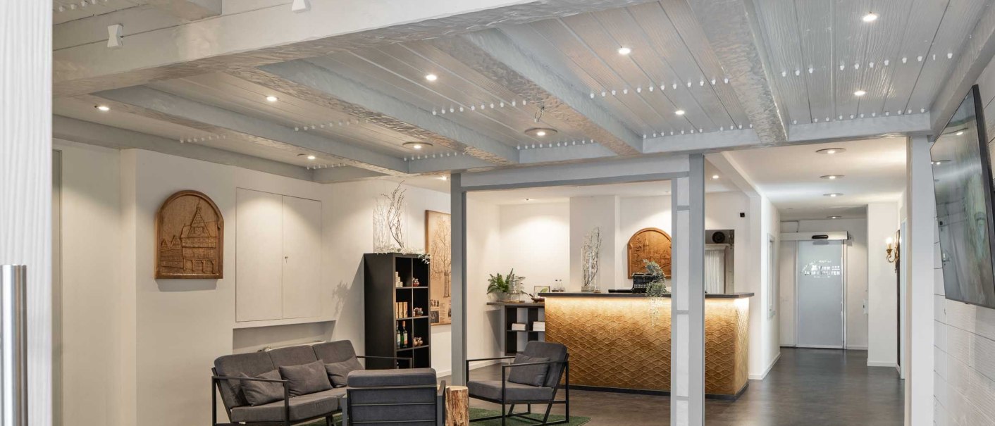 Moderne Rezeption mit gemütlichem Sitzbereich, Holzmöbeln und dekorativer Decke. Helle Beleuchtung und Pflanzen schaffen eine einladende Atmosphäre., © Flairhotel Vier Jahreszeiten