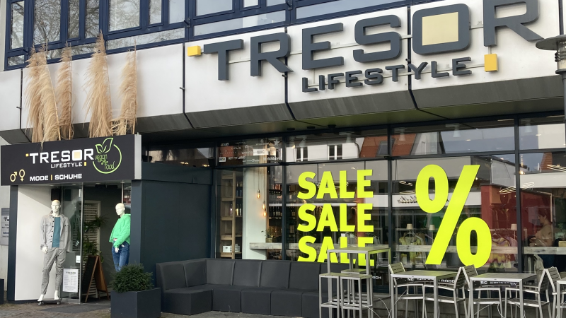 Geschäftseingang von 'Tresor Lifestyle' mit großen Fenstern. Gelbe 'Sale'-Schriftzüge und Sitzgelegenheiten vor dem Laden., © L. Besemer
