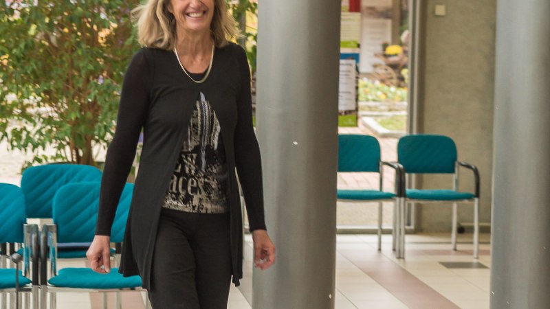 Eine Frau in schwarzer Kleidung geht lächelnd durch einen Raum mit blauen Stühlen und Pflanzen im Hintergrund. Eine Frau in schwarzer Kleidung geht lächelnd durch einen Raum mit blauen Stühlen und Pflanzen im Hintergrund.