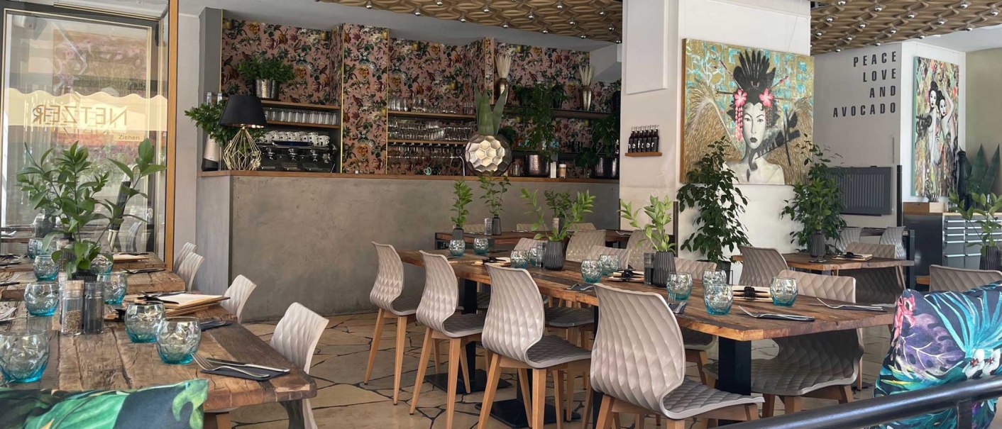 Stilvoll eingerichtetes Restaurant mit Holztischen, modernen Stühlen, Pflanzen und Kunstwerken. Schriftzug "Peace Love and Avocado" an der Wand., © SMG