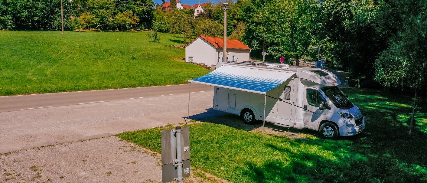 Wohnmobil mit ausgefahrener Markise auf einem Stellplatz neben einer Stra&szlig;e, umgeben von gr&uuml;ner Wiese und B&auml;umen, im Hintergrund H&auml;user., &copy; SMG, Thomas Niederm&uuml;ller