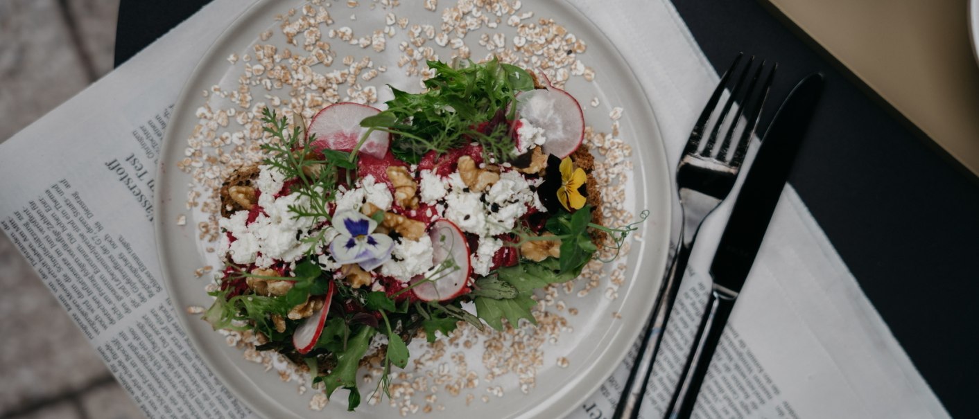 Ein dekorativer Salat mit Radieschen, Nüssen und essbaren Blüten auf einem Teller, serviert auf einer Zeitung., © Fritz, Stuttgart