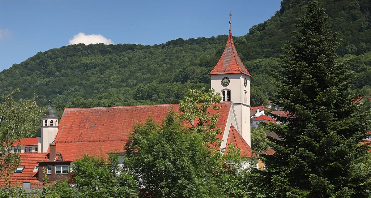Die katholische Kirche St. Bernhard mit rotem Dach und Turm, umgeben von Bäumen, vor einem bewaldeten Hügel unter blauem Himmel., © Katholisches Pfarramt Heubach Die katholische Kirche St. Bernhard mit rotem Dach und Turm, umgeben von Bäumen, vor einem bewaldeten Hügel unter blauem Himmel., © Katholisches Pfarramt Heubach
