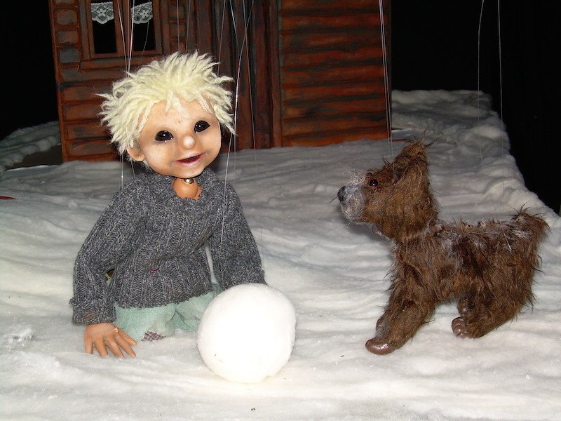 Eine Puppe mit blonden Haaren und grauem Pullover kniet im Schnee neben einem kleinen Hund vor einem Holzhaus., © Zaubermühle Merklingen Eine Puppe mit blonden Haaren und grauem Pullover kniet im Schnee neben einem kleinen Hund vor einem Holzhaus., © Zaubermühle Merklingen