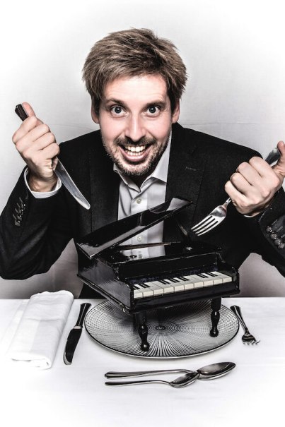 Ein Mann in Anzug hält Messer und Gabel über einem kleinen schwarzen Klavier auf einem Teller. Er lächelt breit., © Kulturhaus Schwanen