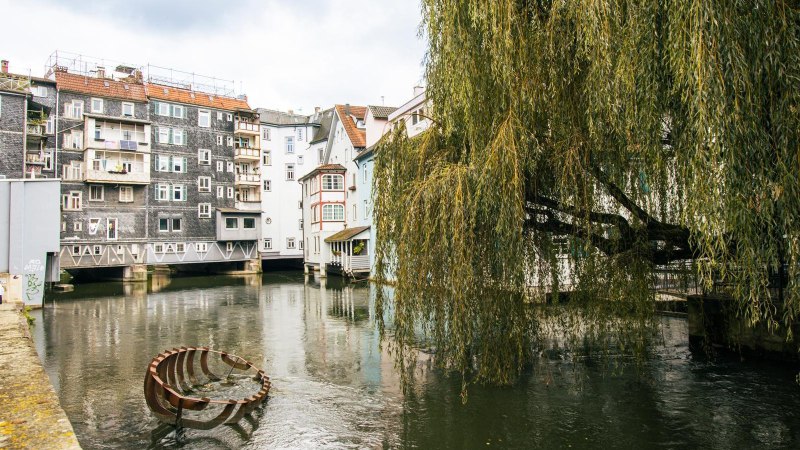 Ein malerischer Fluss flie&szlig;t durch Esslingen, ges&auml;umt von Fachwerkh&auml;usern und einer gro&szlig;en Trauerweide. Ein Kunstwerk ragt aus dem Wasser., &copy; SMG, Sarah Schmid