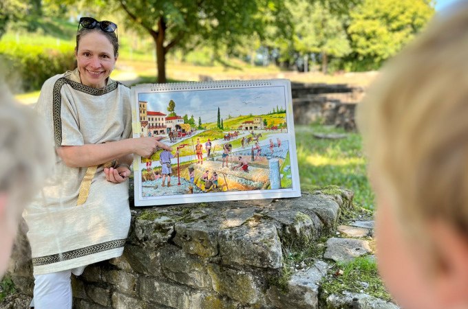 Eine Frau in r&ouml;mischer Kleidung zeigt Kindern ein Bild mit einer r&ouml;mischen Szene. Sie sitzt auf einer Steinmauer im Freien., &copy; Stadt N&uuml;rtingen