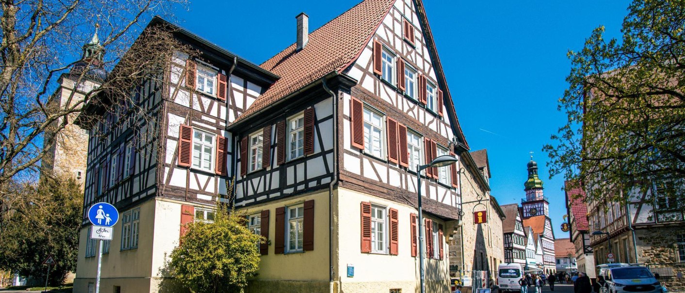 Fachwerkhaus in Kirchheim unter Teck, umgeben von Bäumen und einem Kirchturm im Hintergrund. Menschen und Autos beleben die Straße., © Stuttgart-Marketing GmbH, Sarah Schmid