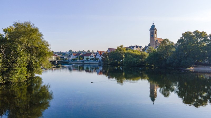 Blick über den Neckar auf eine Kirche und Häuser, umgeben von Bäumen, die sich im Wasser spiegeln., © Stadt Nürtingen