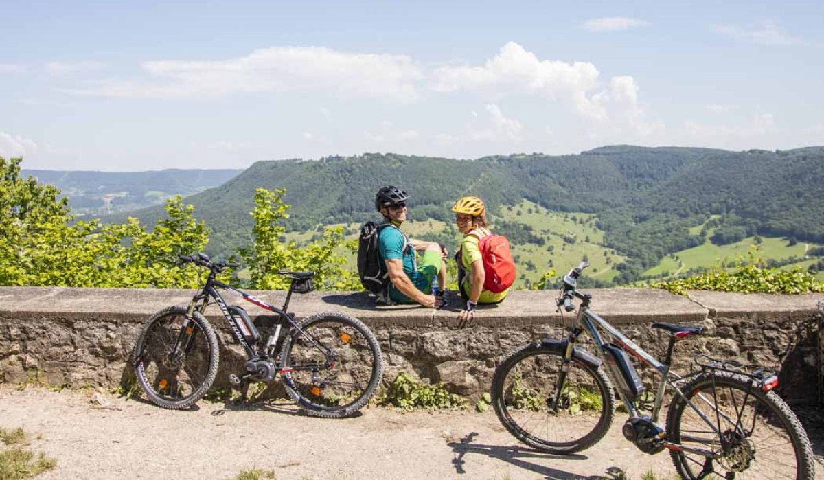 Zwei Radfahrer sitzen auf einer Mauer und genießen den Ausblick auf die grüne Hügellandschaft. Zwei Fahrräder stehen daneben., © Schwäbische Alb Tourismusverband e.V. Zwei Radfahrer sitzen auf einer Mauer und genießen den Ausblick auf die grüne Hügellandschaft. Zwei Fahrräder stehen daneben., © Schwäbische Alb Tourismusverband e.V.