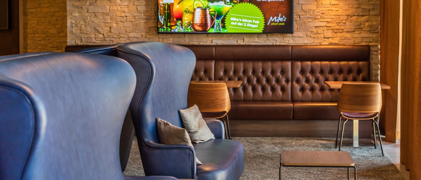 Moderne Hotelbar mit blauen Sesseln, braunen Ledersofas und einem Bildschirm mit Werbung für Mike's Urban Pub an der Wand., © Hilton Garden Inn Stuttgart Neckarpark Moderne Hotelbar mit blauen Sesseln, braunen Ledersofas und einem Bildschirm mit Werbung für Mike's Urban Pub an der Wand., © Hilton Garden Inn Stuttgart Neckarpark