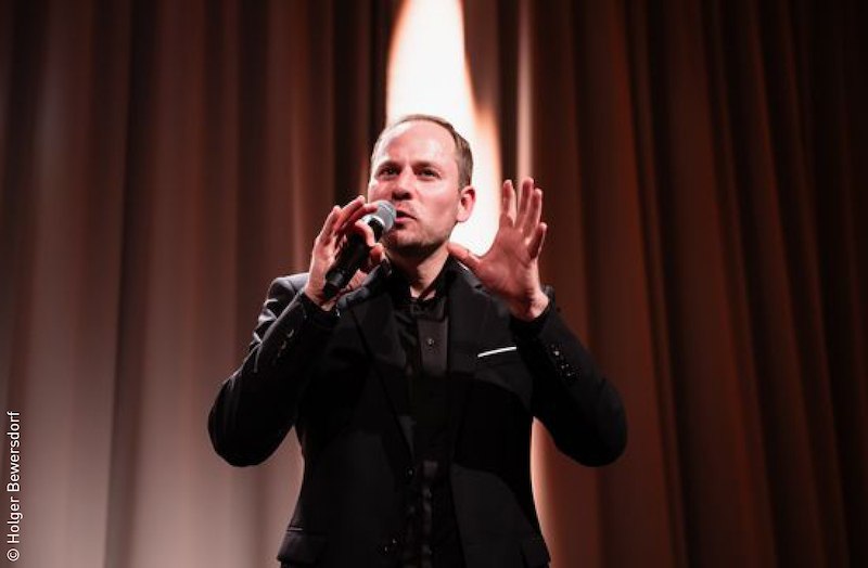 Ein Mann in schwarzem Anzug spricht leidenschaftlich in ein Mikrofon auf einer Bühne mit braunem Vorhang., © Stadt Kornwestheim Kulturmanagement Ein Mann in schwarzem Anzug spricht leidenschaftlich in ein Mikrofon auf einer Bühne mit braunem Vorhang., © Stadt Kornwestheim Kulturmanagement