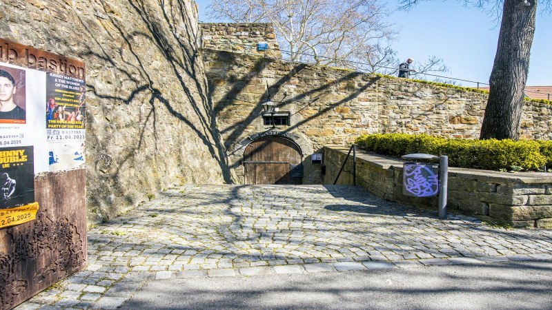 Eingang zum Club Bastion in Kirchheim unter Teck. Alte Steinmauer, Holztor, Plakate an der Wand. Kopfsteinpflasterweg, Baum und Büsche im Vordergrund., © SMG, Sarah Schmid Eingang zum Club Bastion in Kirchheim unter Teck. Alte Steinmauer, Holztor, Plakate an der Wand. Kopfsteinpflasterweg, Baum und Büsche im Vordergrund., © SMG, Sarah Schmid