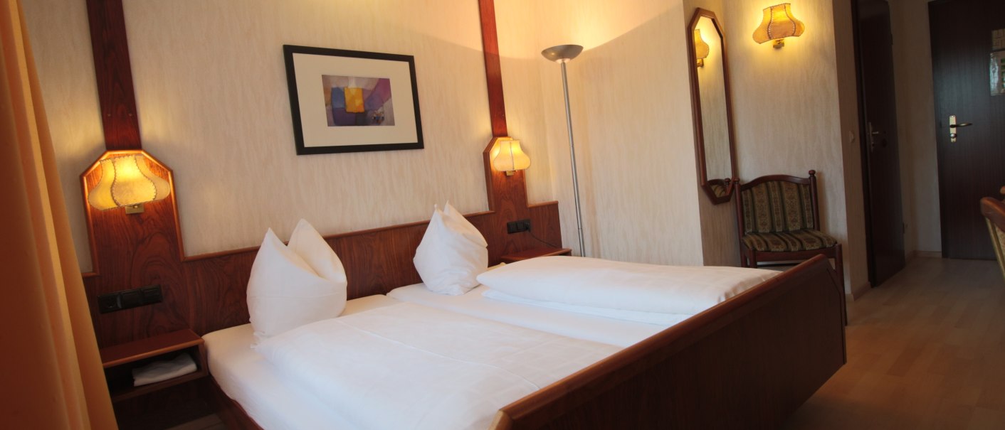 Gemütliches Doppelzimmer mit Holzmöbeln, großem Bett, Wandlampen und Bild an der Wand. Helle, warme Beleuchtung und ein Stuhl in der Ecke., © TOMAS Gemütliches Doppelzimmer mit Holzmöbeln, großem Bett, Wandlampen und Bild an der Wand. Helle, warme Beleuchtung und ein Stuhl in der Ecke., © TOMAS