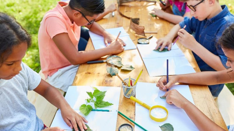 Kinder sitzen an einem Holztisch im Freien und basteln mit Blättern und Papier. Sie verwenden Stifte und Lupen für ihre kreativen Projekte., © AdobeStock_Robert_Kneschke Kinder sitzen an einem Holztisch im Freien und basteln mit Blättern und Papier. Sie verwenden Stifte und Lupen für ihre kreativen Projekte., © AdobeStock_Robert_Kneschke