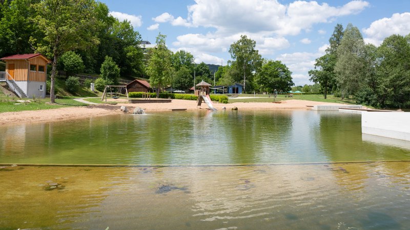 Das Kocherfreibad in Künzelsau zeigt einen Sandstrand, einen Spielplatz mit Rutsche und Schaukeln, umgeben von Bäumen und einem klaren Himmel., © Olivier Schniepp, Foto Linke GmbH Das Kocherfreibad in Künzelsau zeigt einen Sandstrand, einen Spielplatz mit Rutsche und Schaukeln, umgeben von Bäumen und einem klaren Himmel., © Olivier Schniepp, Foto Linke GmbH