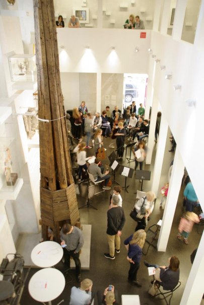 Menschen versammeln sich in einem modernen Raum mit hohen Decken. Ein gro&szlig;es Holzobjekt steht zentral. Einige Personen sitzen, andere stehen und unterhalten sich., &copy; Ludwigsburg Museum